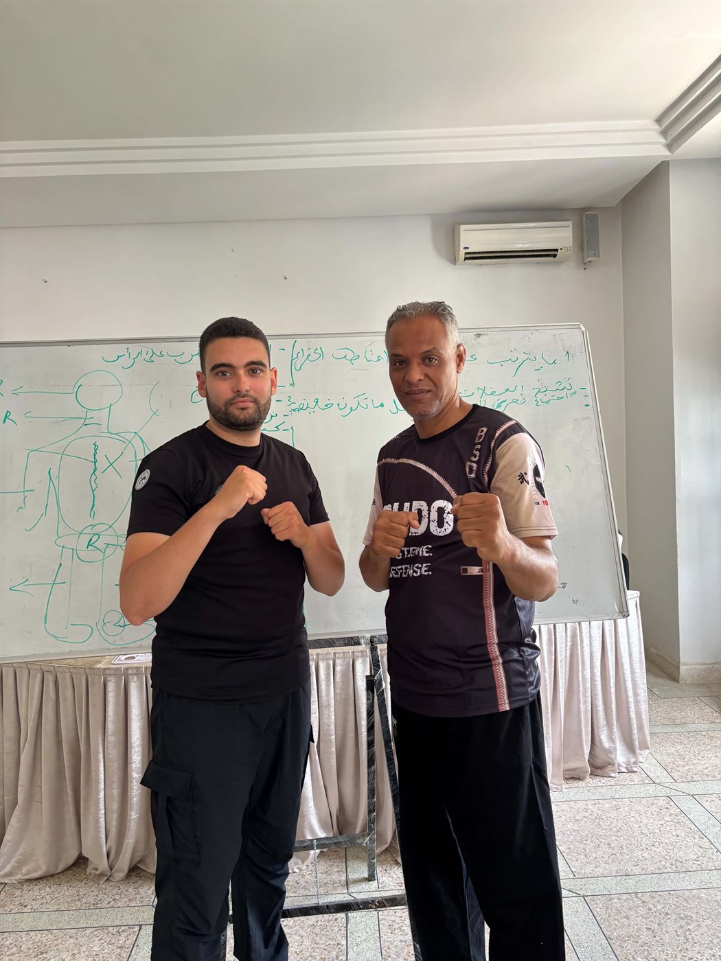 Two martial arts instructors standing together in front of a training whiteboard